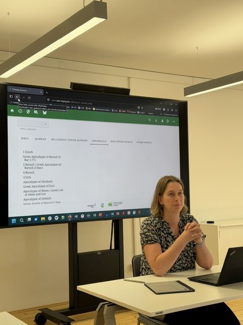 Im Hintergrund befindet sich ein riesiger Monitor, darauf Text. Davor sitzt eine Frau an einem Tisch mit aufgeklapptem Monitor und Pad sowie Stift. Sie schaut an der Kamera vorbei.